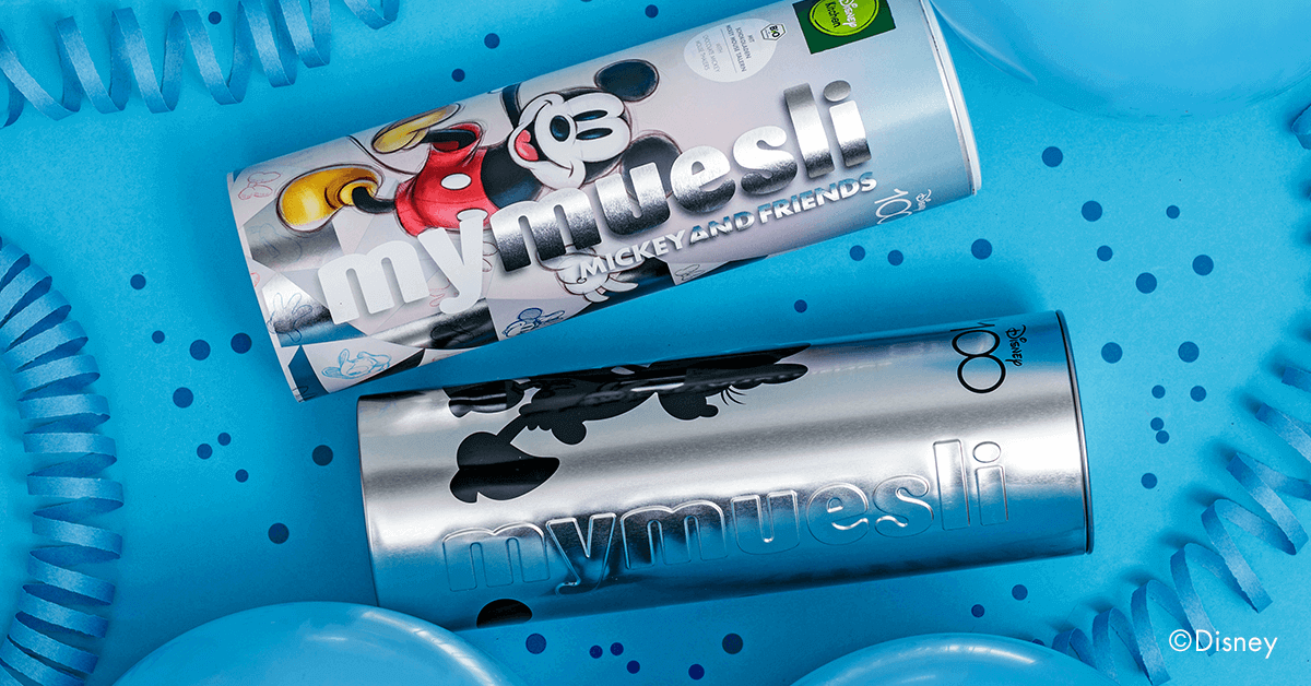 Disney Fan Paket zum Jubilaeum mit Vorratsdose in Silber und Mickey and Friends Muesli auf festlichem Hintergrund mit Konfetti und Papierschlangen