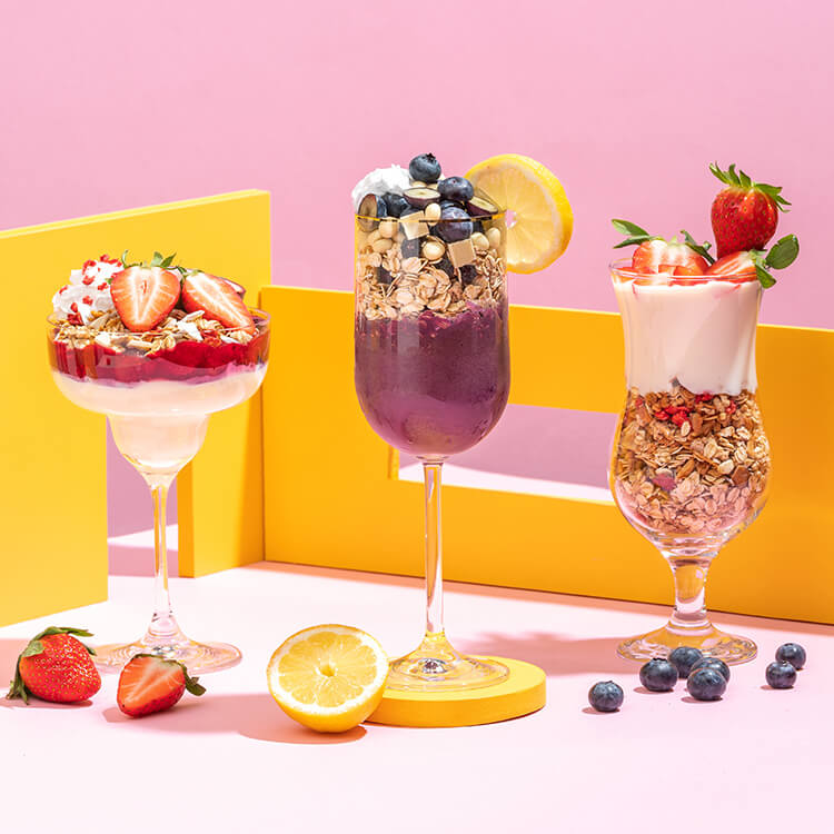 article1a-muesli-summer-minis.jpg Drei Cocktailgläser mit Muesli Desserts gefuellt vor sommerlich hellem Hintergrund